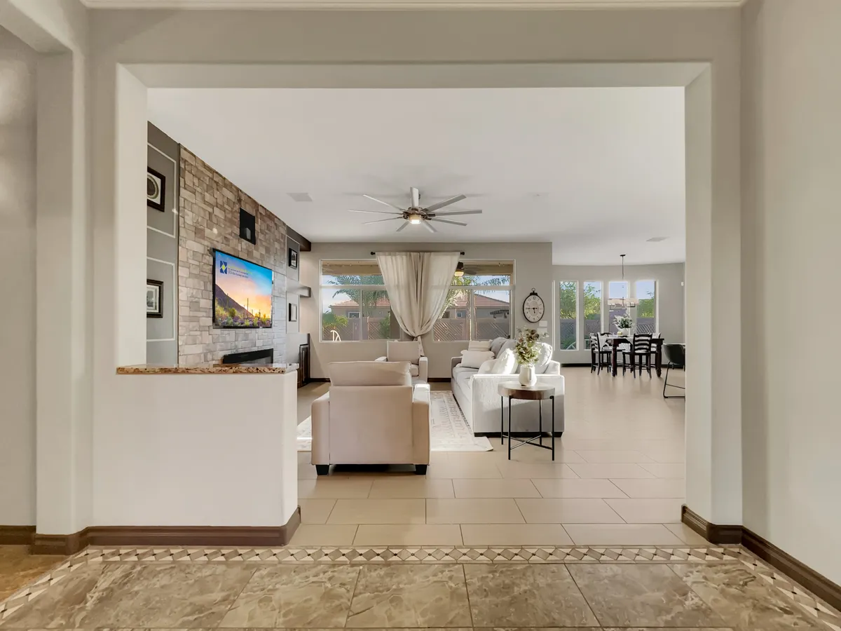 This image captures a spacious living room with a comfortable seating area, a TV mounted on a striking stone wall, and large windows that allow natural light to flood the space. The background reveals an adjacent dining area, subtly connecting the living and dining spaces.