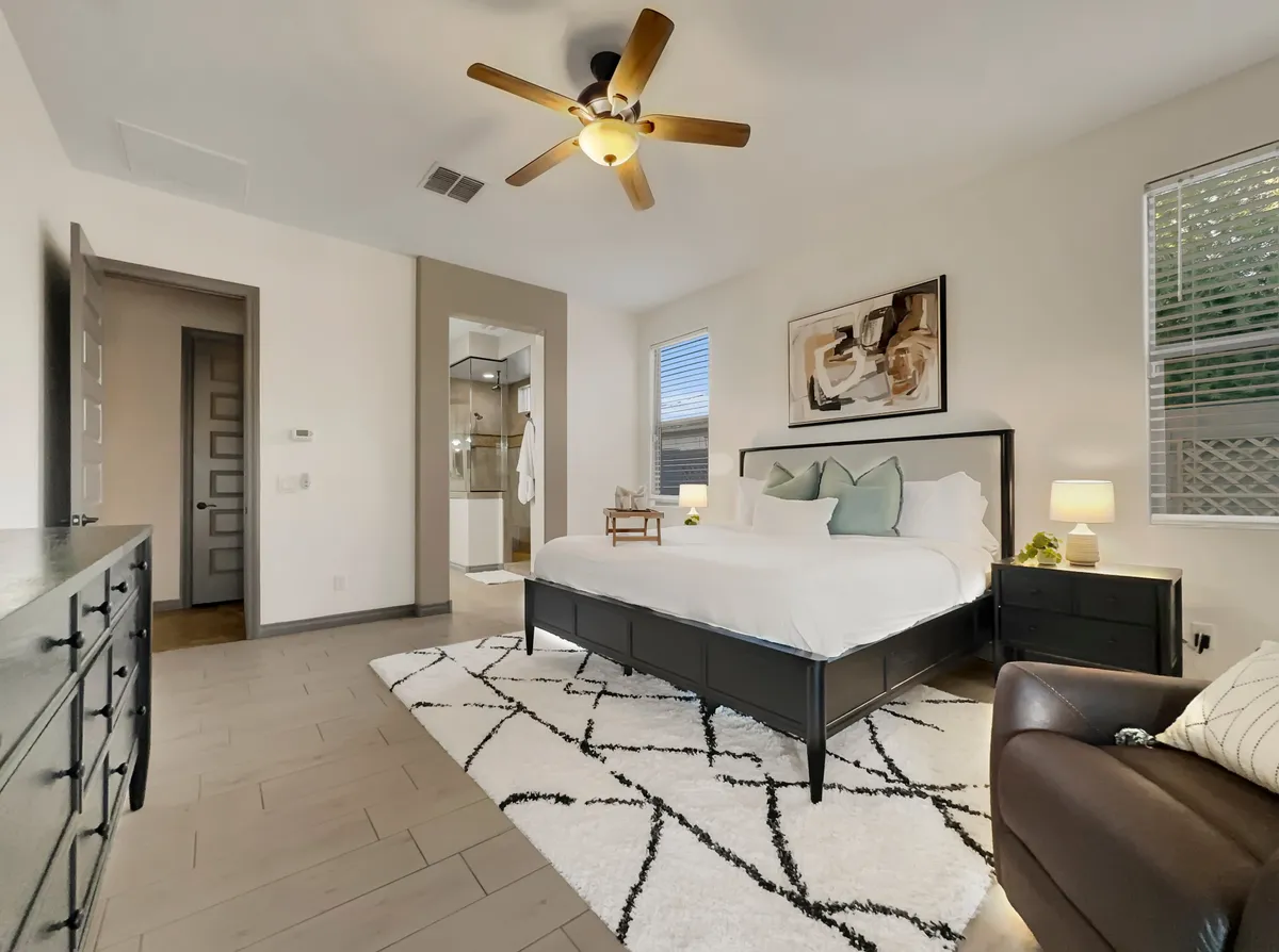 A serene master bedroom with a king-sized bed adorned with decorative pillows, soft rugs underfoot, tasteful wall art, and nightstands with glowing lamps. Natural light fills the space, creating a peaceful retreat ideal for rest and relaxation.