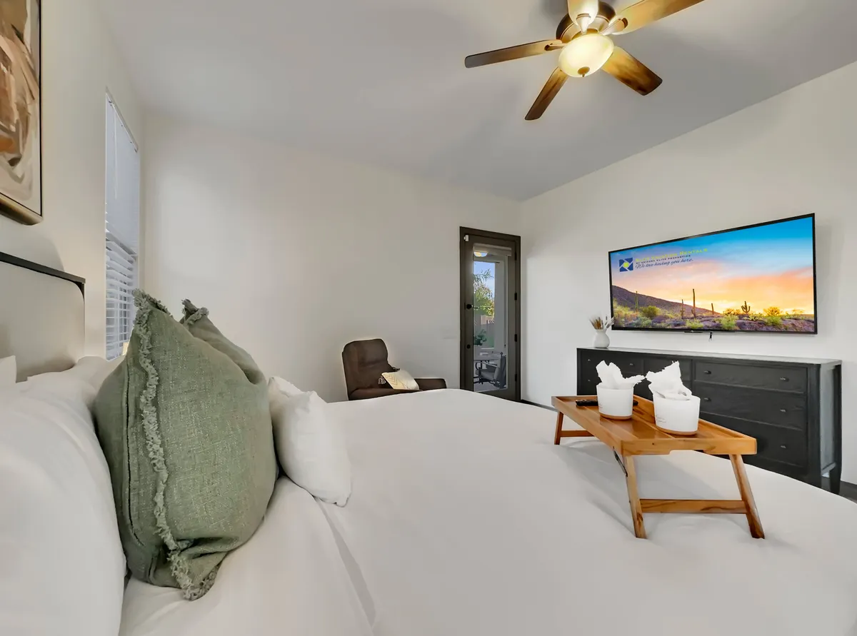 A cozy queen bedroom featuring a large bed with crisp white linens, a modern TV mounted on the wall, a wooden side table with decorative items, and a comfortable armchair by the window. The space is bright and inviting, ideal for private relaxation or entertainment.