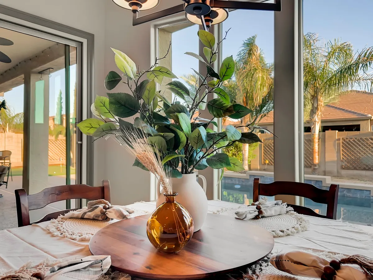 The interior dining area features a wooden table, decorative vase with plants, and large windows that offer glimpses of the outdoor pool and garden. Natural light floods the space, creating a seamless connection between indoor relaxation and outdoor amenities.