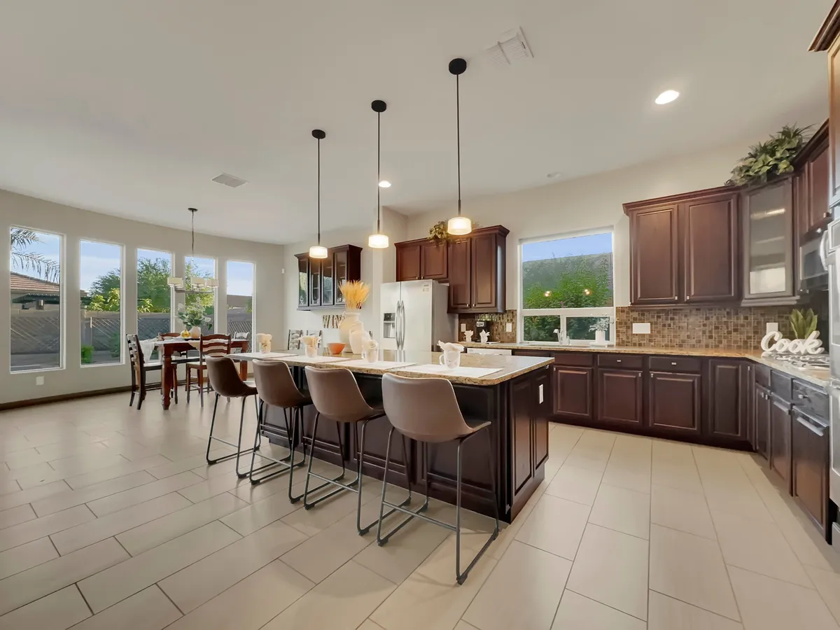 This scene features a modern kitchen with a prominent island and bar stools, flowing seamlessly into a dining area with a well-set table. Large windows brighten the space, while the layout encourages socializing and shared activities.