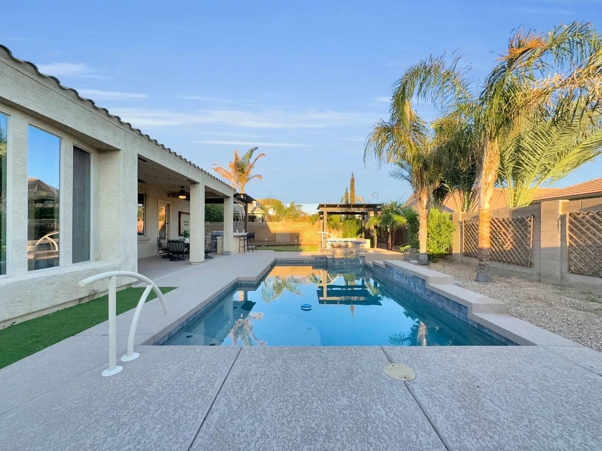 A tranquil backyard view featuring a swimming pool flanked by palm trees, a covered patio area, and inviting lounge chairs. The sun casts a soft glow over the scene, ideal for sunbathing or enjoying the shade.