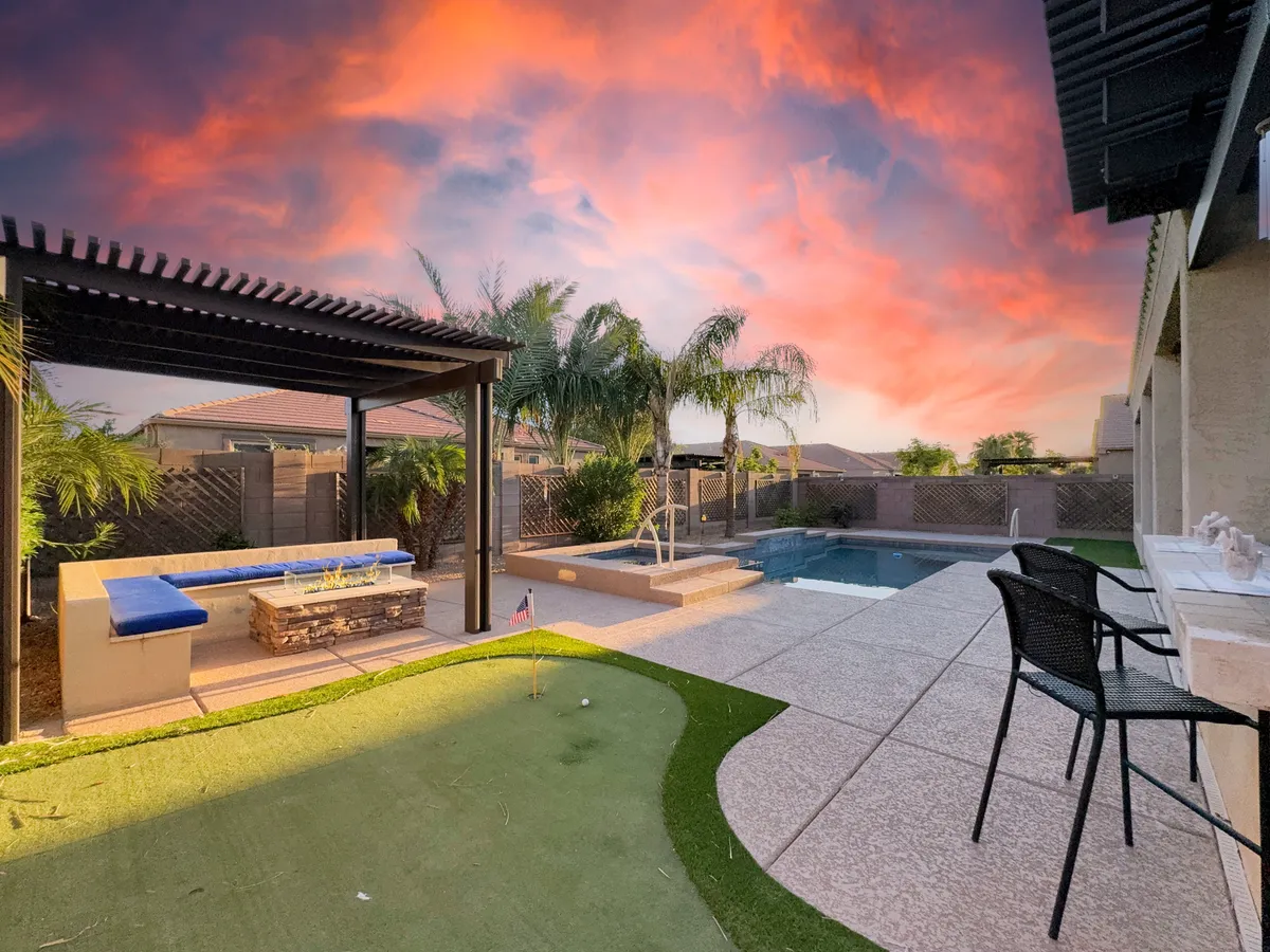 A vibrant exterior scene featuring a putting green, swimming pool, ample outdoor seating, palm trees, and a colorful sunset sky. The space feels secure and inviting, ideal for both people and pets to enjoy outdoor activities together.