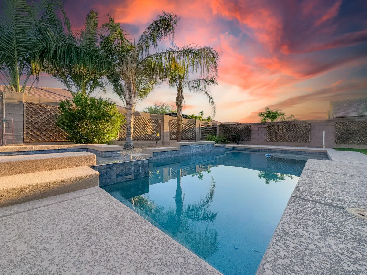 A stunning view of the backyard oasis at sunset, featuring a resort-style heated swimming pool flanked by tropical palm trees and a colorful sky. The inviting water and spacious poolside area suggest plenty of room for group activities, relaxation, and evening gatherings.