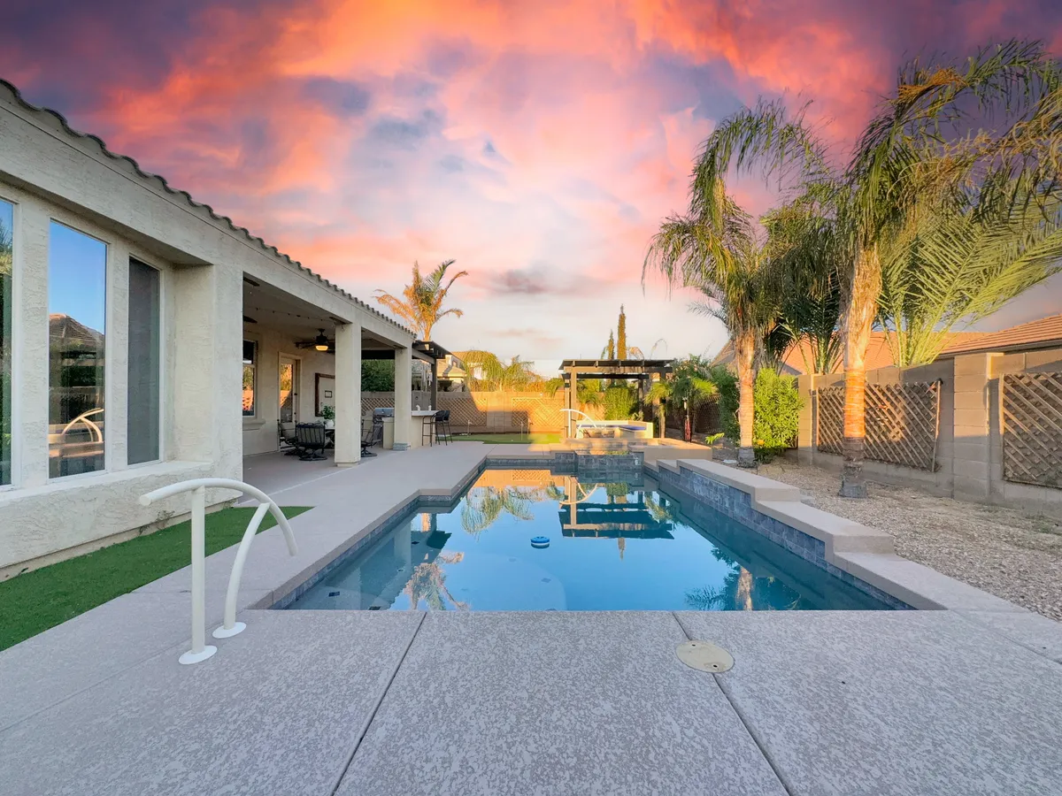 A picturesque backyard scene with a swimming pool surrounded by palm trees, a covered patio for shade, and a colorful sky at sunset. The mood is joyful and relaxed, ideal for evening swims or quiet family gatherings.